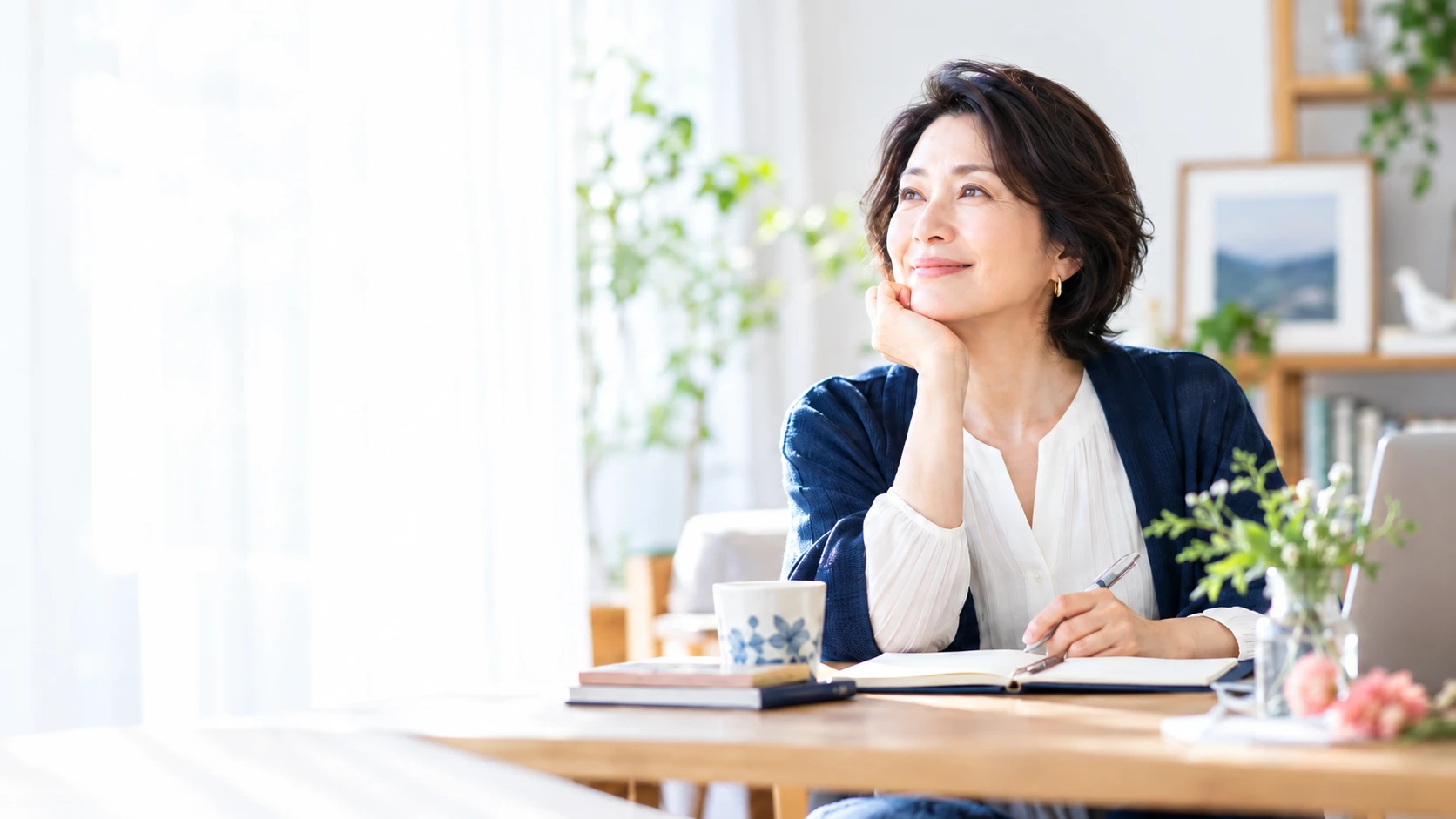 窓辺のテーブルでノートを広げ、穏やかに外を見る50代の女性